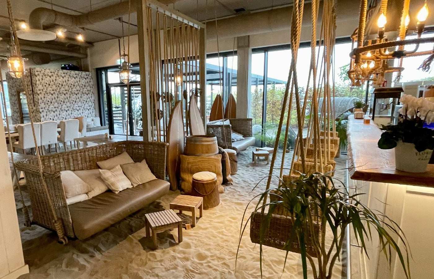 Lounge mit Hängesesseln, Sand und Surfbrettern hinter großen Fenstern