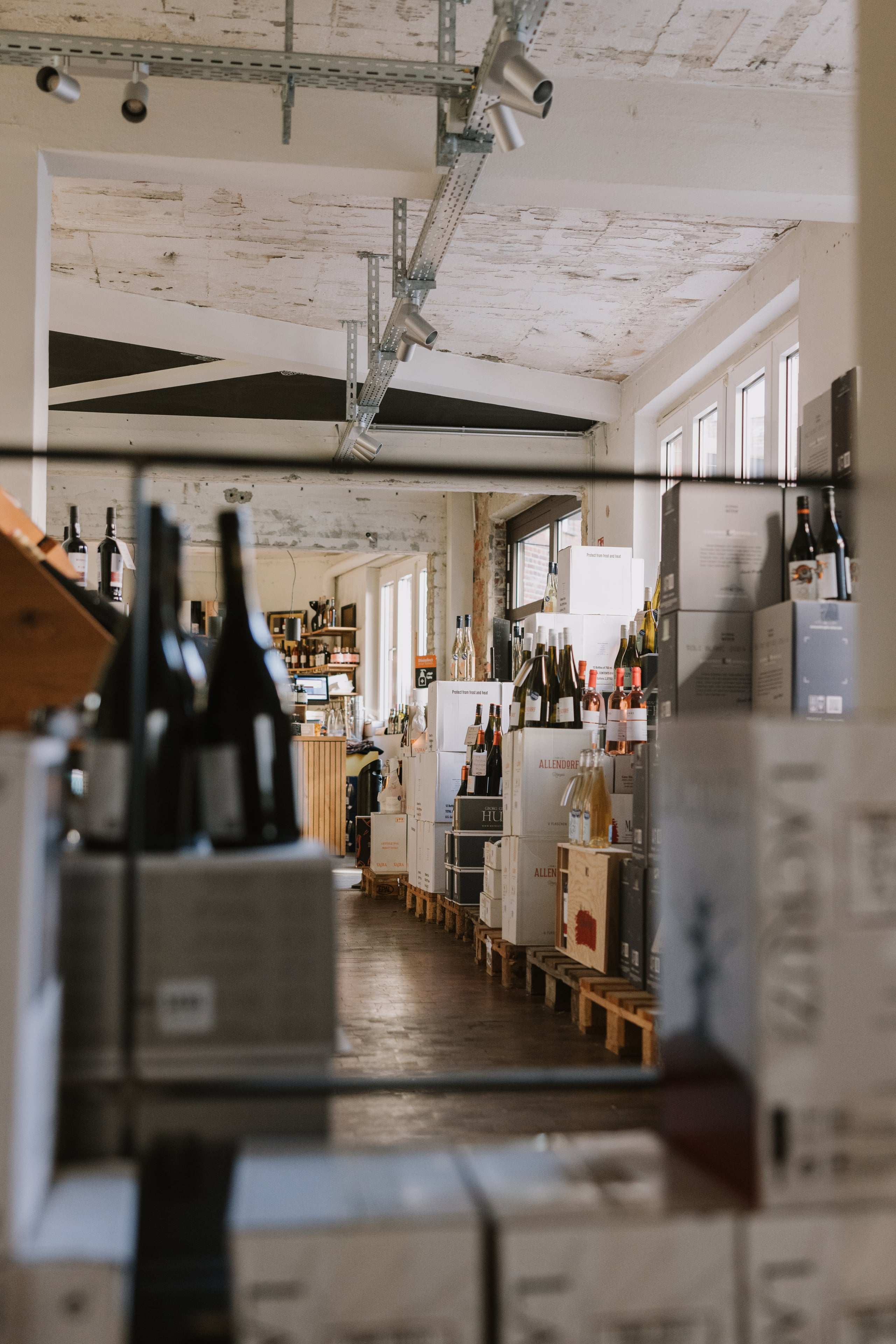 Der lange Raum des Weinlagers mit Weinkartons und Flaschen auf Paletten, durch eine Flaschenreihe im Vordergrund gesehen