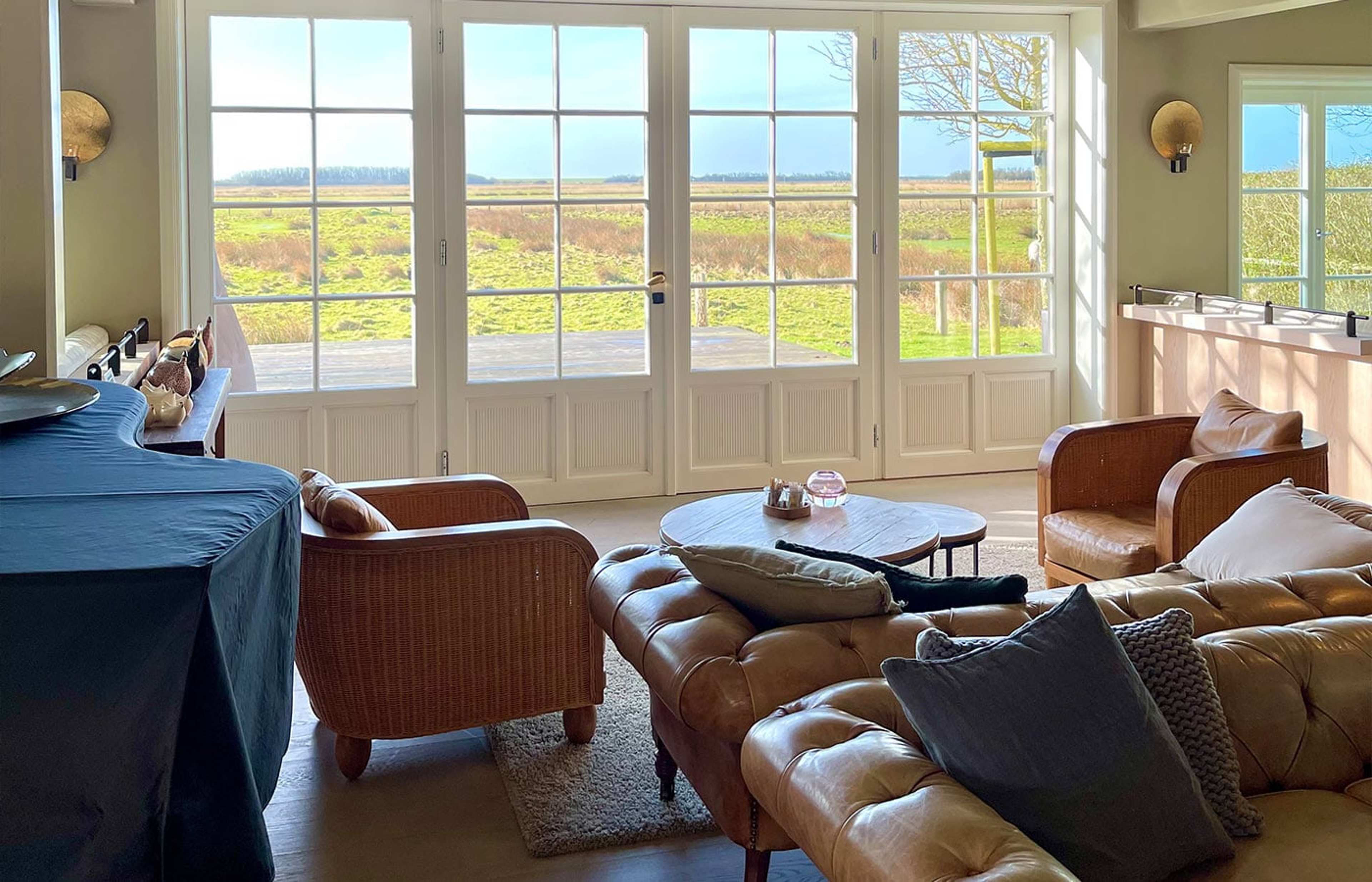 Heller Salon mit Ledersofas und Sesseln, durch die Sprossenfenster der Blick in die Marsch