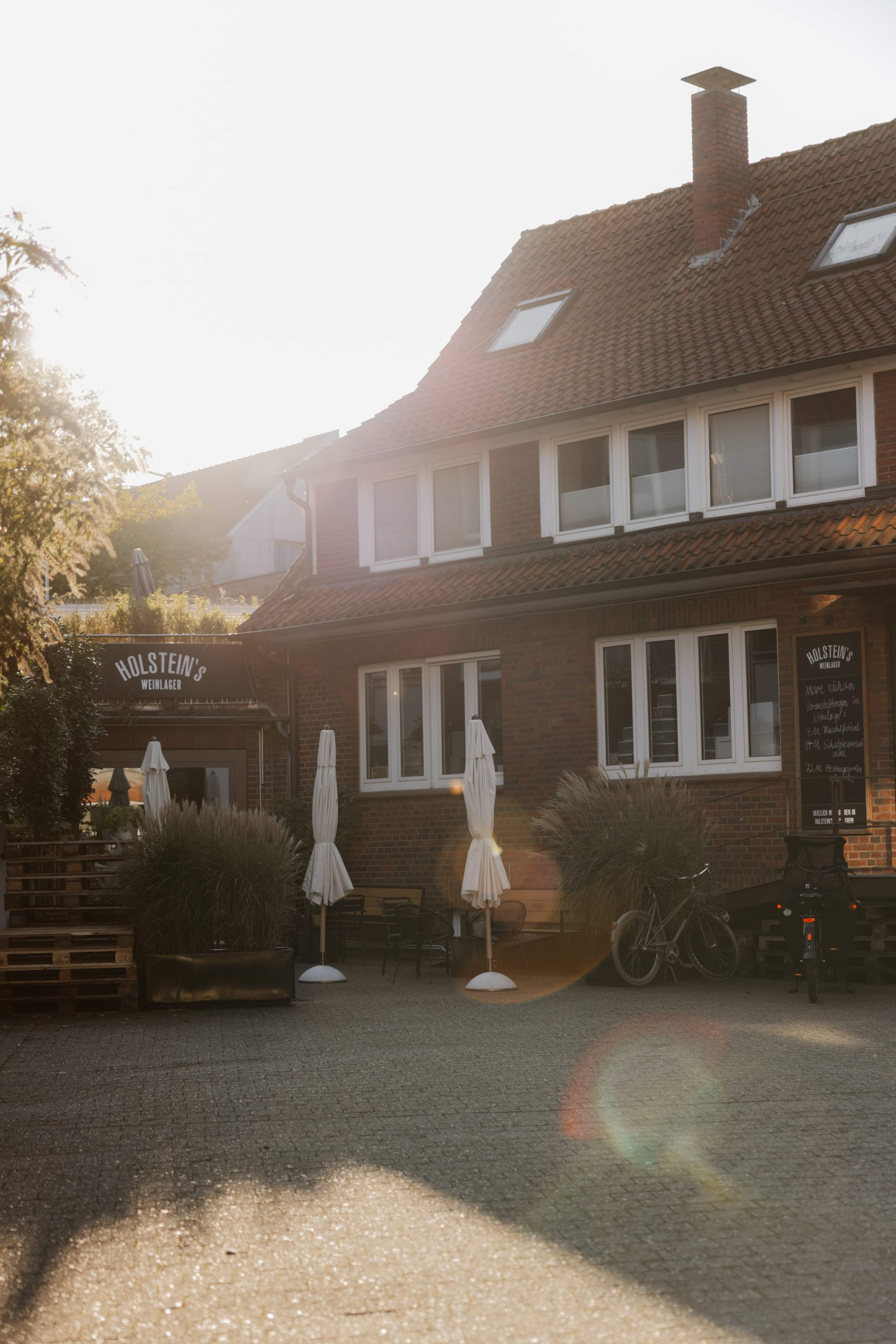 Das Backsteinhaus des Weinlagers mit Holstein's-Schild, Sonnenschirmen und Hof im Gegenlicht der Abendsonne