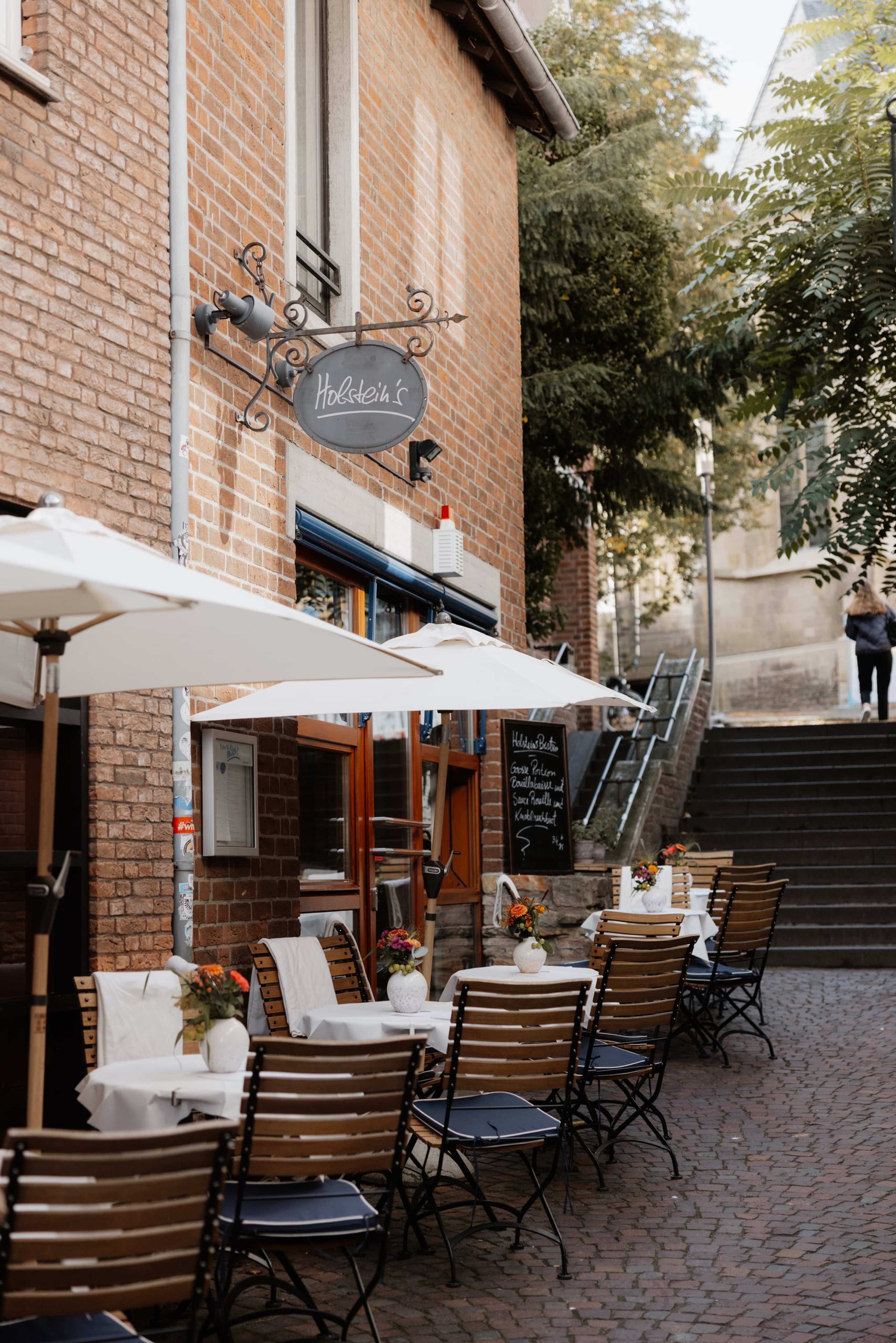 Terrasse von Holsteins Bistro mit weißen Schirmen und Tischen, darüber das Schild Holstein's