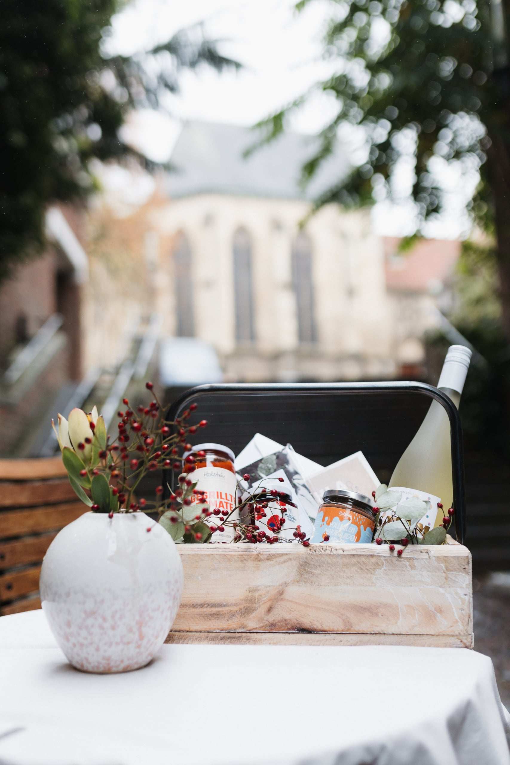 Präsent in einer Holzkiste mit Aufstrichen, Weißwein und Karten, daneben eine Vase mit Hagebutten und Protea, im Hintergrund eine Kirche
