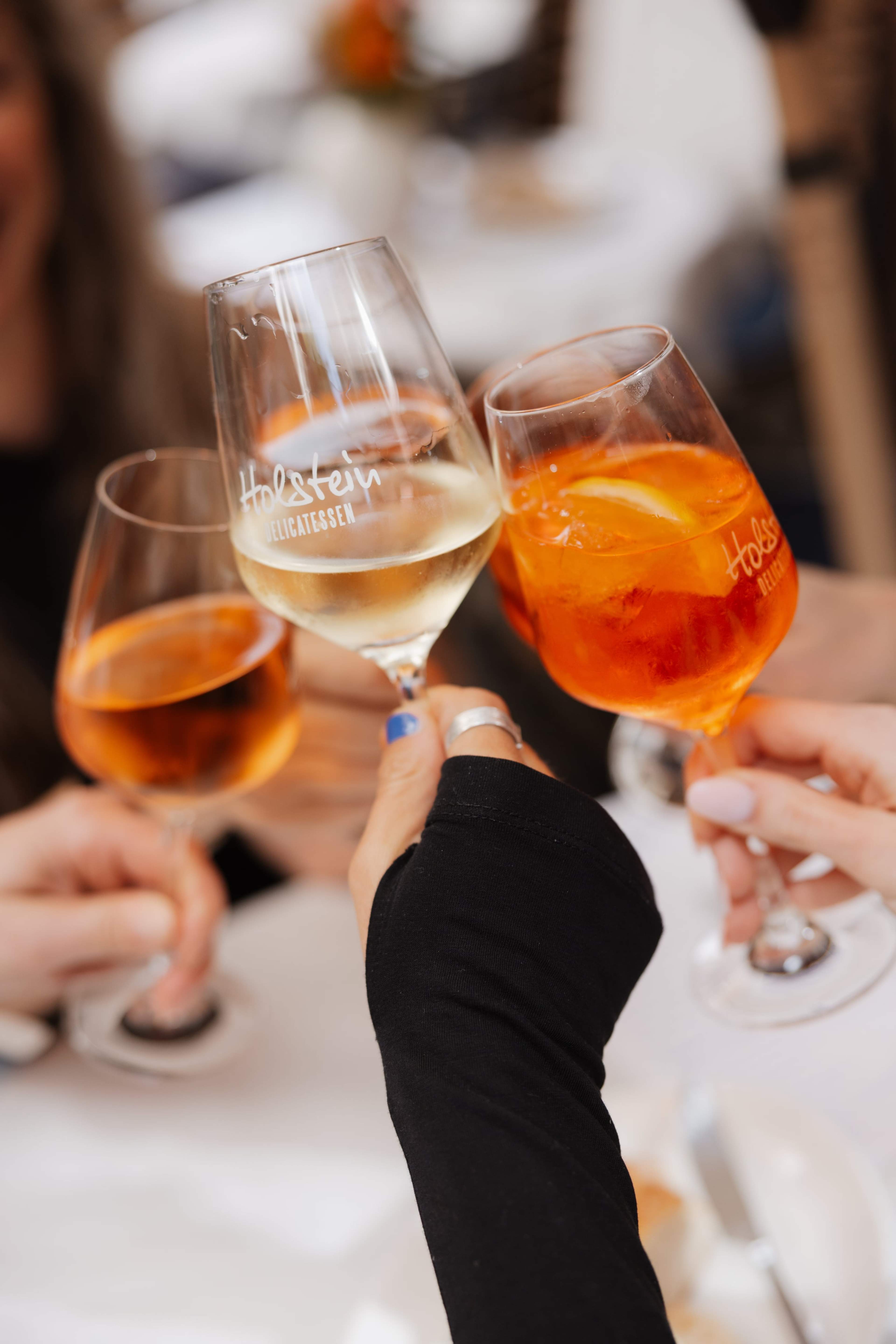 Hände stoßen mit Weißwein und Spritz in Gläsern mit Holstein-Gravur an