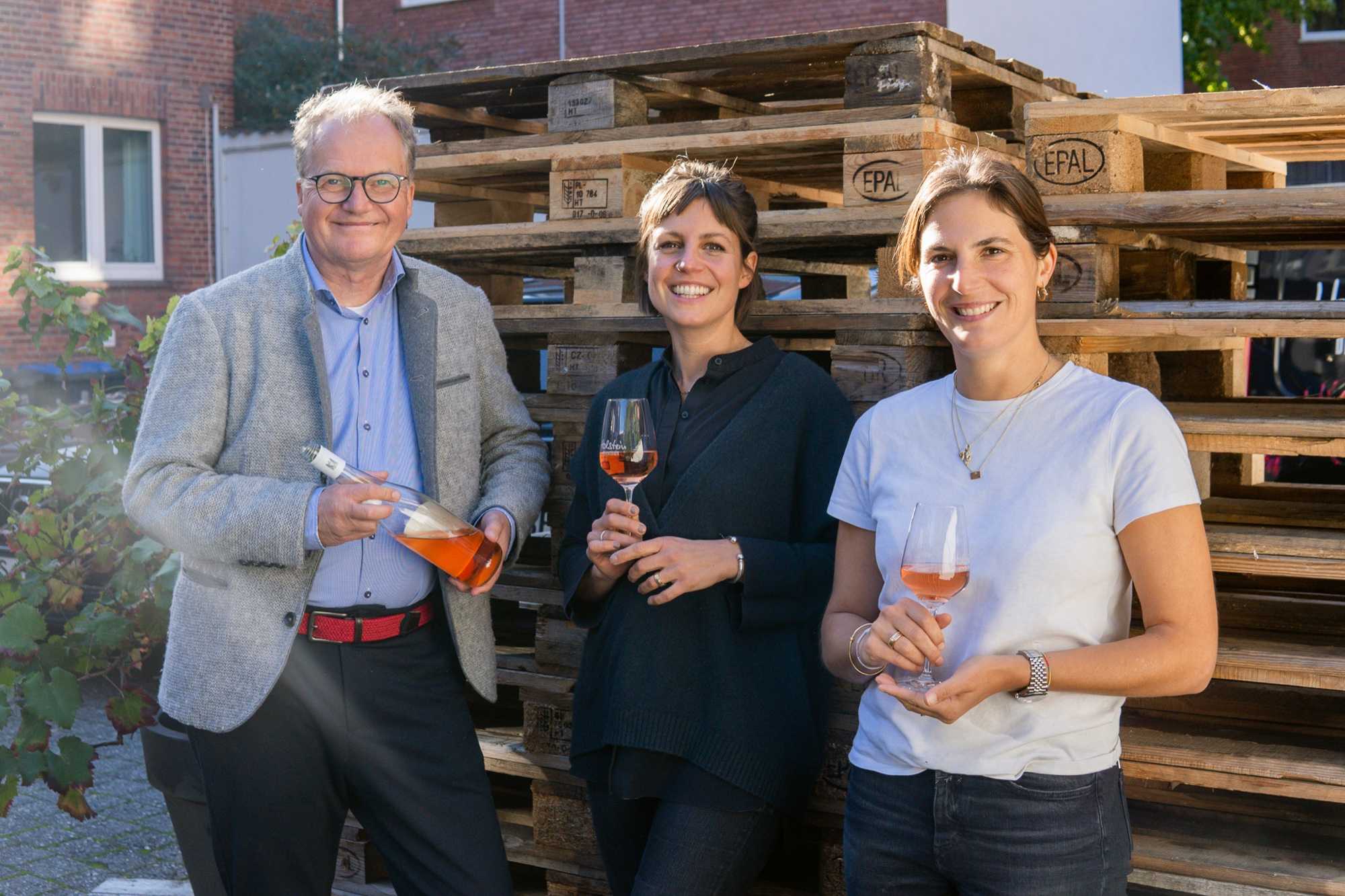 Klaus Helmrich mit einer Flasche Roséwein, neben ihm zwei Frauen mit je einem Glas Rosé, vor gestapelten Holzpaletten und einem Backsteinhaus