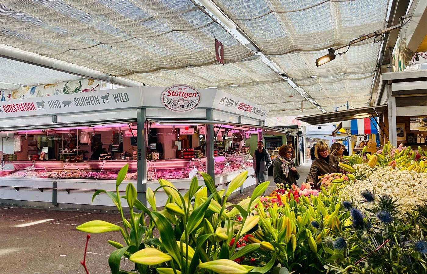 Marktstände unter einem Sonnensegel, vorne Blumen