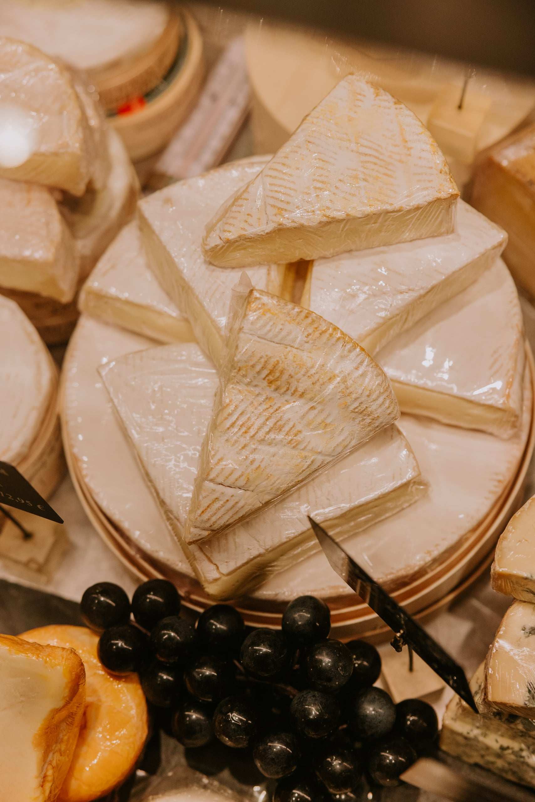 Angeschnittene Weichkäse-Laibe und Brie-Stücke in der Käsetheke, davor dunkle Trauben