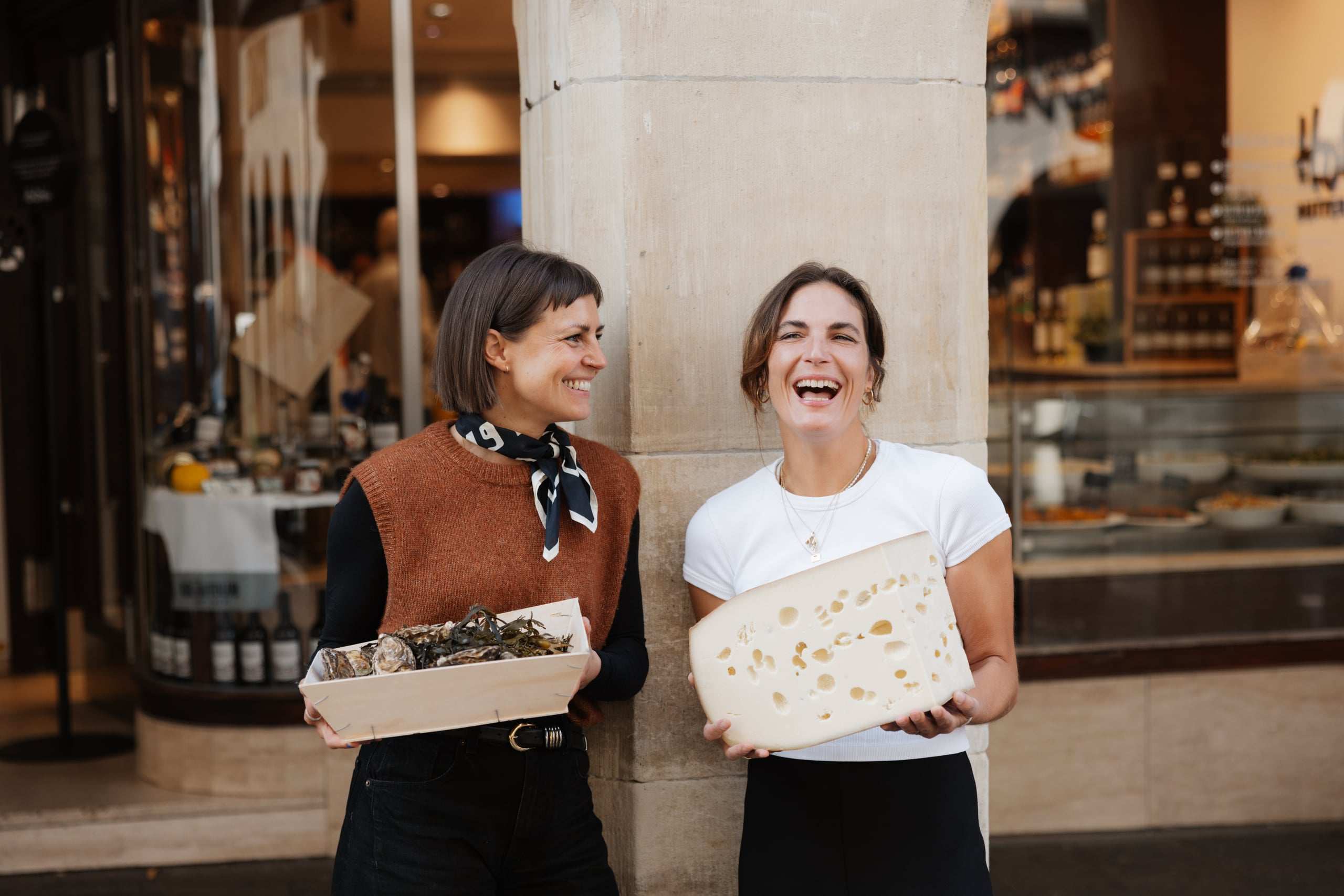 Judith und Sophie Helmrich lachen herzlich vor dem Schaufenster der Butterhandlung, mit einer Kiste Austern und einem großen Stück Käse