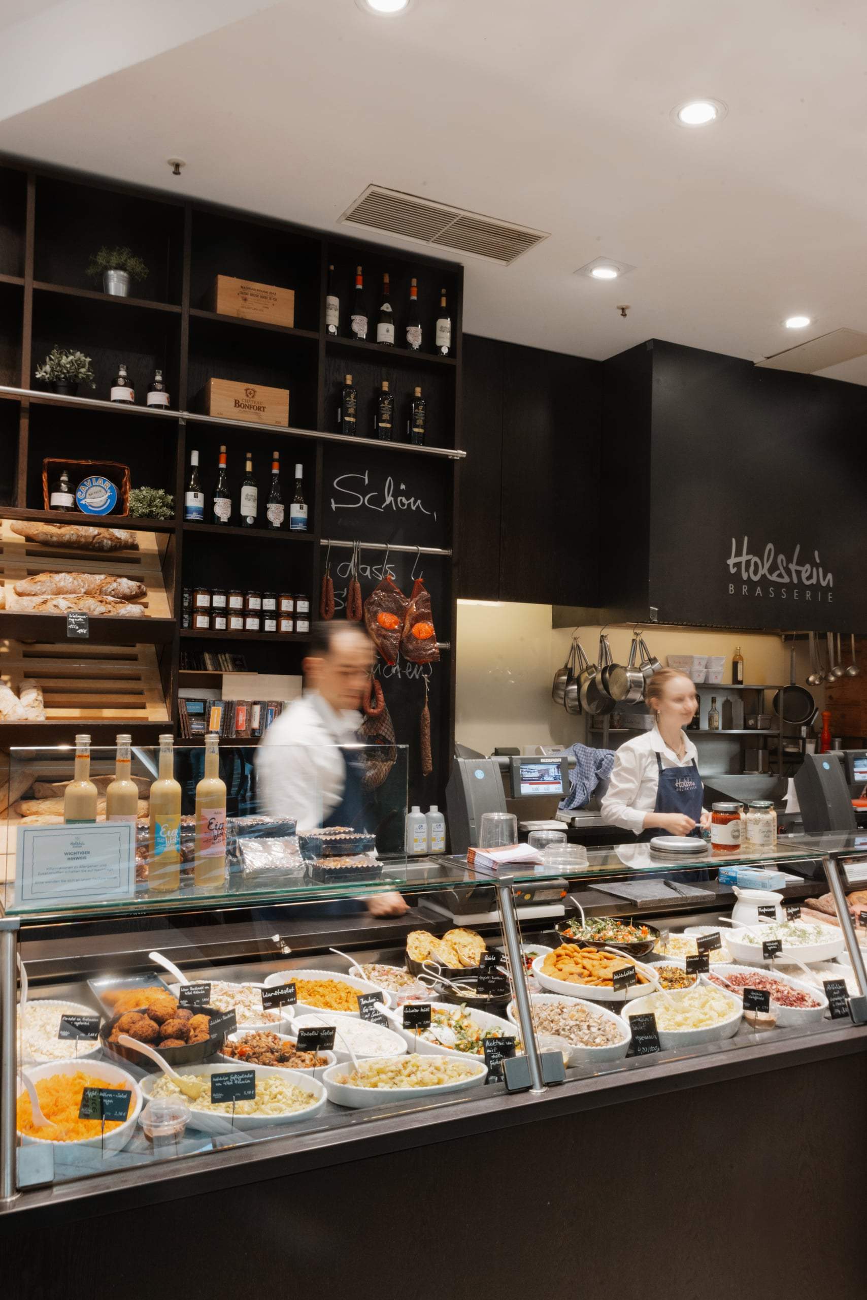 Deli-Theke mit zwei Mitarbeitenden, an der Wand der Schriftzug Holstein Brasserie