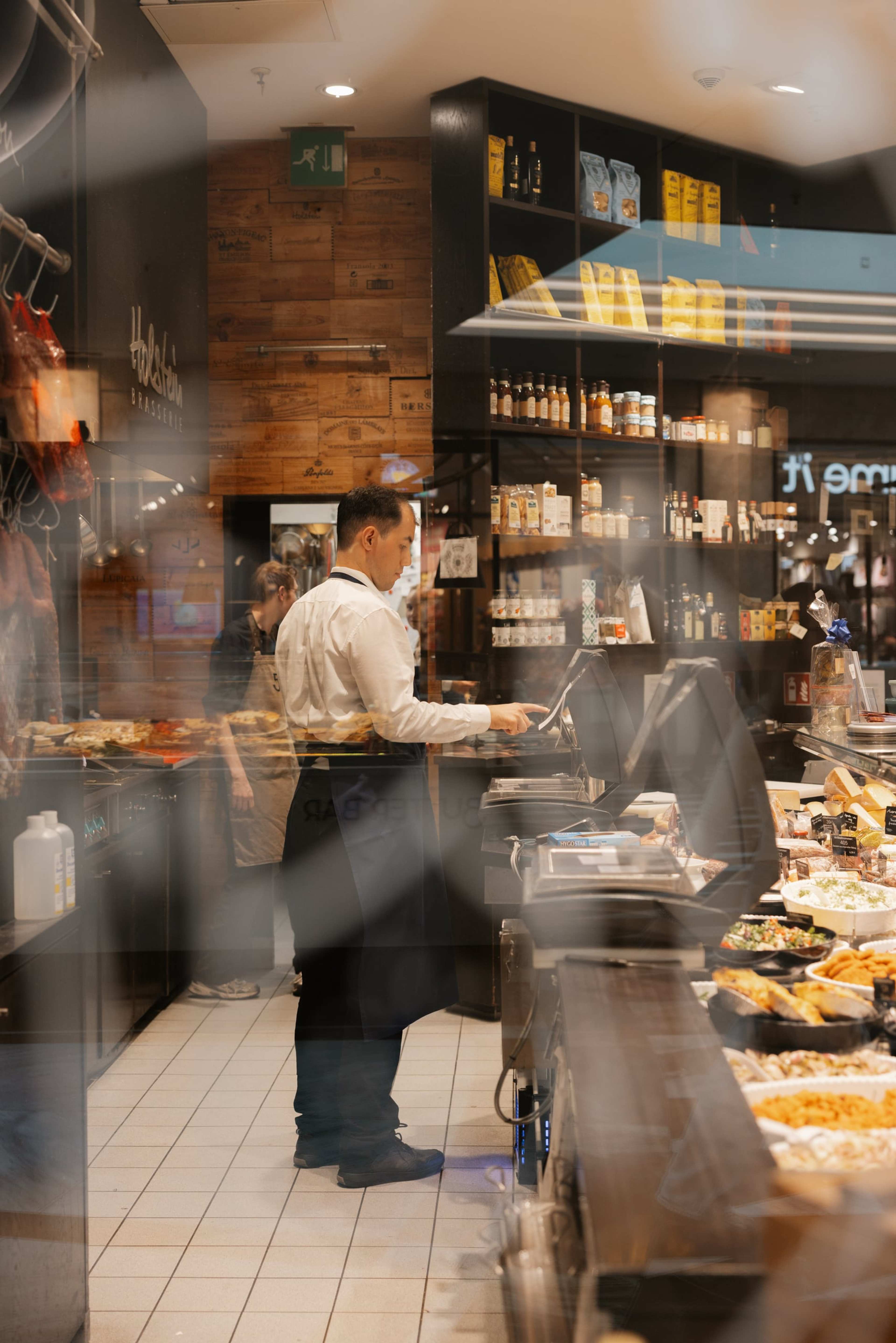 Blick durch die Scheibe in das Deli: ein Mitarbeiter in weißem Hemd hinter der Theke, dahinter Regale